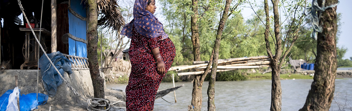 A woman stands on the eroded edge of her home, looking out over a rising riverbank in a coastal area, highlighting vulnerability to climate change.