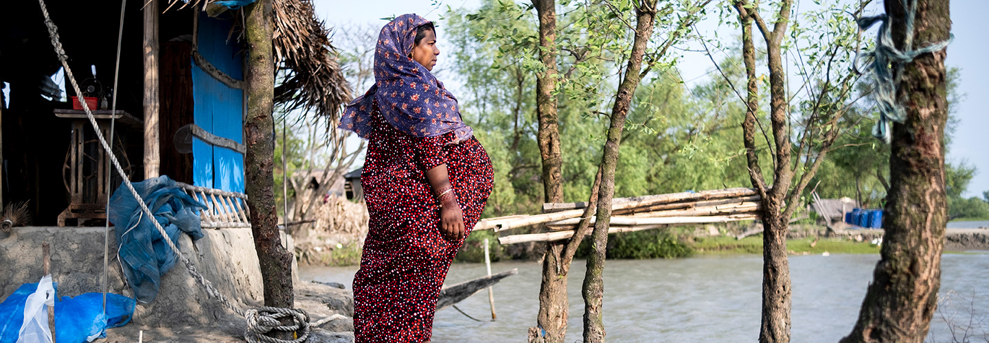 A pregnant woman in a red dress and headscarf stands outside a thatched-roof home near a flooded area, gazing into the distance, with trees and a makeshift wooden bridge nearby.