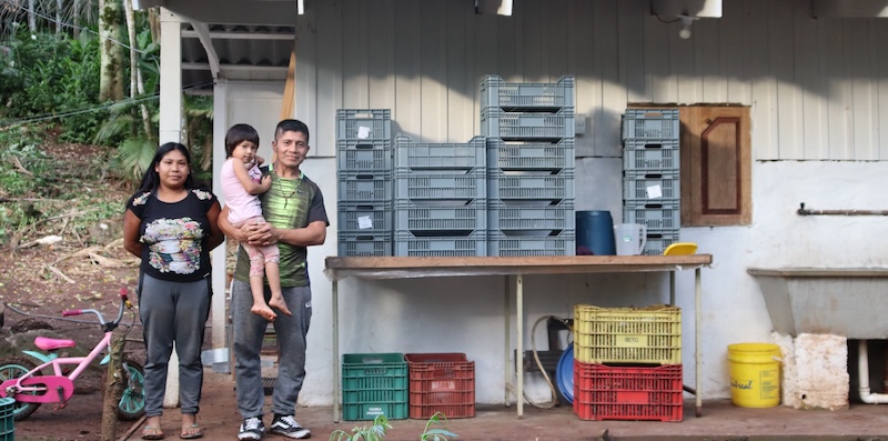 A Guaraní family of three stand next to an açaí processing unit.