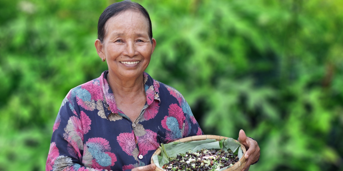 A smiling woman holds a woven basket filled with harvested seeds or plants, set against a lush green background.