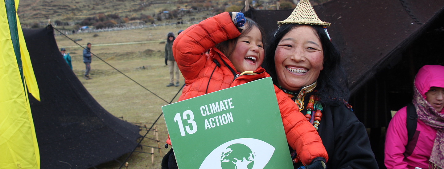 A smiling woman in traditional Bhutanese attire holds a laughing child and a sign for SDG 13: Climate Action, set against a rural mountainous background.