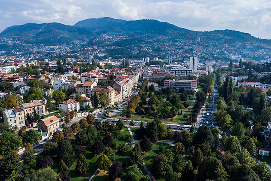  An aerial view of Sarajevo showcasing a mix of urban buildings, lush green parks, and tree-lined streets, with a backdrop of surrounding mountains under a partly cloudy sky.