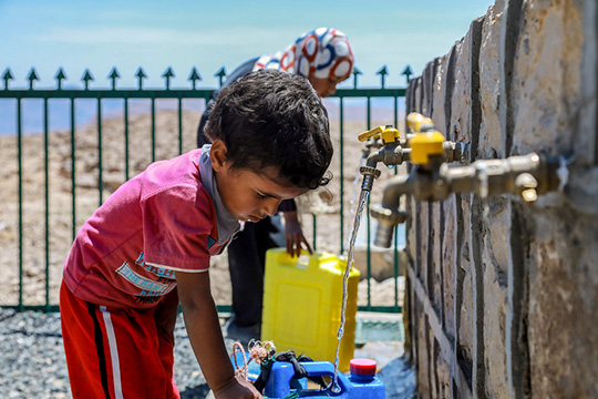 A young boy fills a container with water from a public tap, while a woman in the background carries a yellow jerry can near a fenced area in a dry, arid landscape.