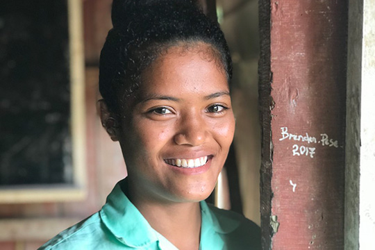  A smiling young woman stands inside a rustic building, leaning against a wooden post.