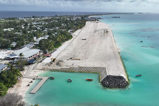 An aerial view of a land reclamation project in Tuvalu, showing a wide sandy area bordered by turquoise waters and a small coastal settlement with buildings and palm trees.