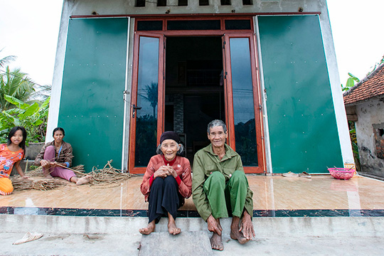  An elderly couple sits on the tiled front step of a green, storm-resilient house, with two younger family members in the background preparing materials.