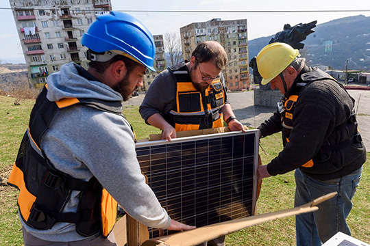  Three workers wearing helmets and safety vests assemble a solar panel outdoors, with apartment buildings and a hillside visible in the background.