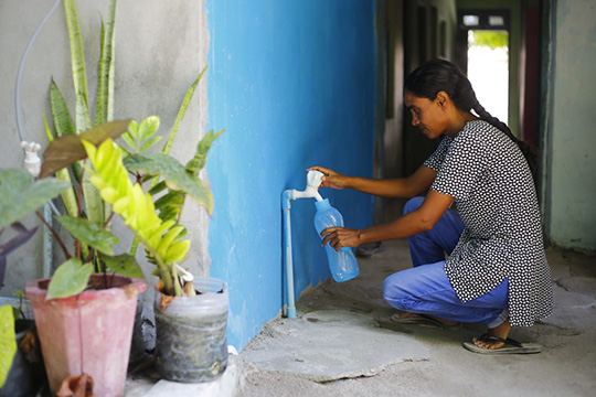 A woman fills a bottle with water from a tap attached to a bright blue wall, with potted plants nearby in a hallway setting.