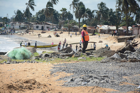 A worker in a reflective vest and helmet stands on a rocky coastline while people and fishing boats are visible along a sandy beach lined with palm trees and small buildings in the background.