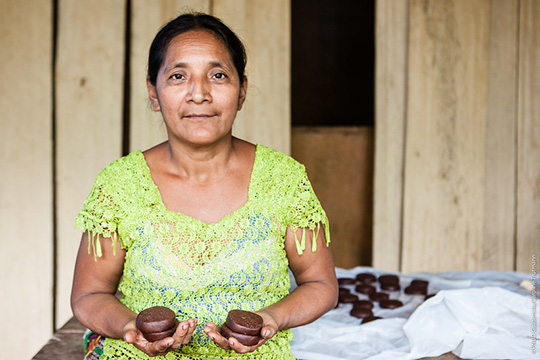 A woman wearing a bright green lace blouse holds cocoa products in her hands, with more cocoa items displayed on a table behind her in a rustic wooden setting.