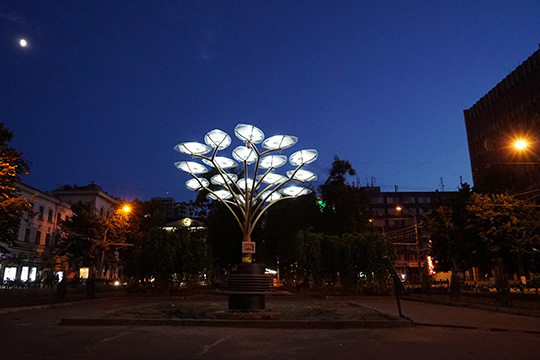  A solar-powered tree-shaped structure with illuminated panels stands in a city square at dusk, surrounded by urban buildings and streetlights.