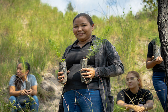  A young woman smiles while holding two saplings in soil bags, with other students sitting and holding saplings in a grassy outdoor area during a reforestation activity.