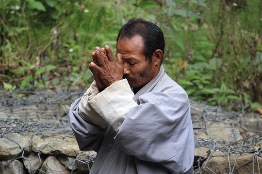  An older man wearing traditional attire holds his hands together in a gesture of prayer or respect, standing outdoors with greenery and stone gabions in the background.