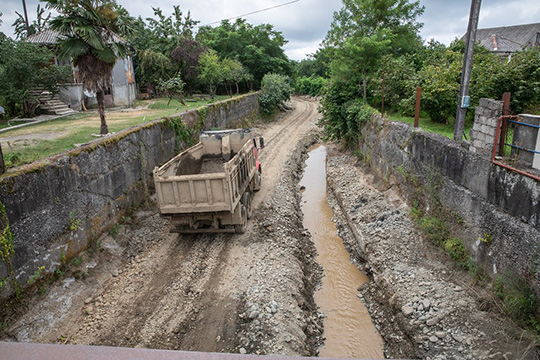 A truck drives through a partially dried riverbed undergoing maintenance, with muddy water in the channel and lush greenery and houses lining the banks.