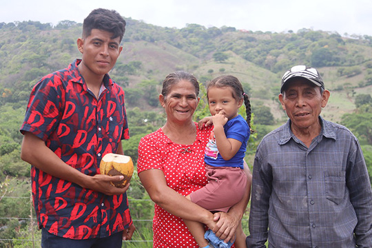 A smiling family of four, including a young man holding a coconut, a woman in a red polka-dot dress holding a small child, and an older man in a cap, stands in front of a green hillside.