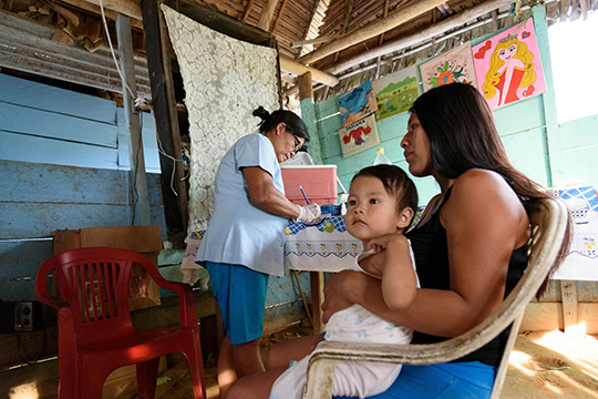 A woman sits holding a young child on her lap while a healthcare worker writes in a notebook inside a modest wooden home decorated with colorful drawings.