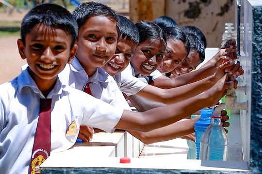 A group of smiling schoolchildren in uniform fill bottles from a row of outdoor water taps at their school.