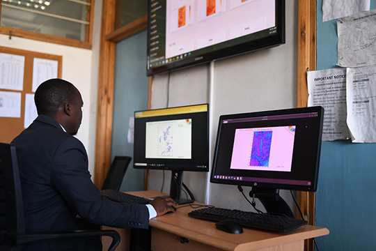 A man sits at a desk analyzing climate data maps displayed on two computer monitors, with additional information projected on a large screen in an office setting.