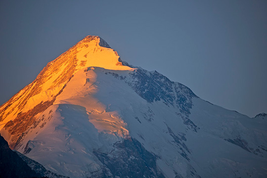 A snow-covered mountain peak glows with golden sunlight against a deep blue sky during sunrise.