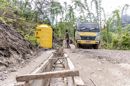 Workers prepare to construct a drainage system, with a yellow water tank and a truck in the background, surrounded by a lush green hillside.