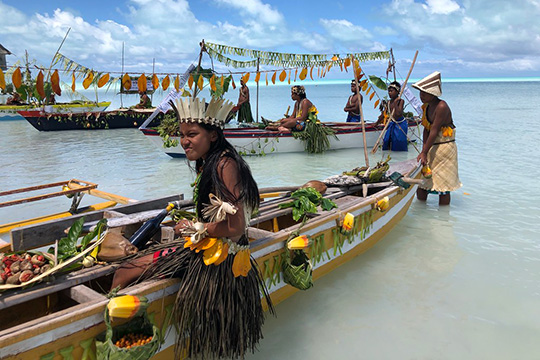 People dressed in traditional attire decorate and display canoes with local produce and leaves in shallow turquoise waters during a cultural celebration on Maiana Island.